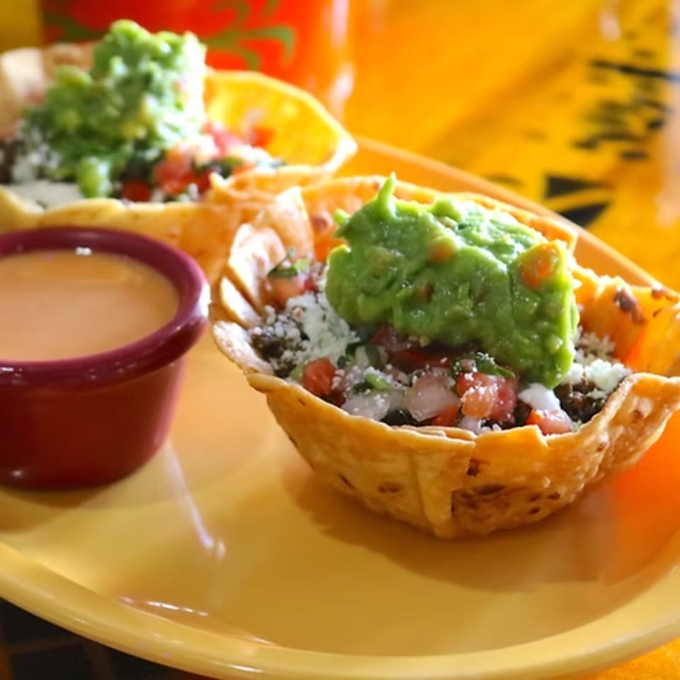 Two crispy taco bowls filled with meat, pico de gallo, cheese, and guacamole, with a side of sauce.