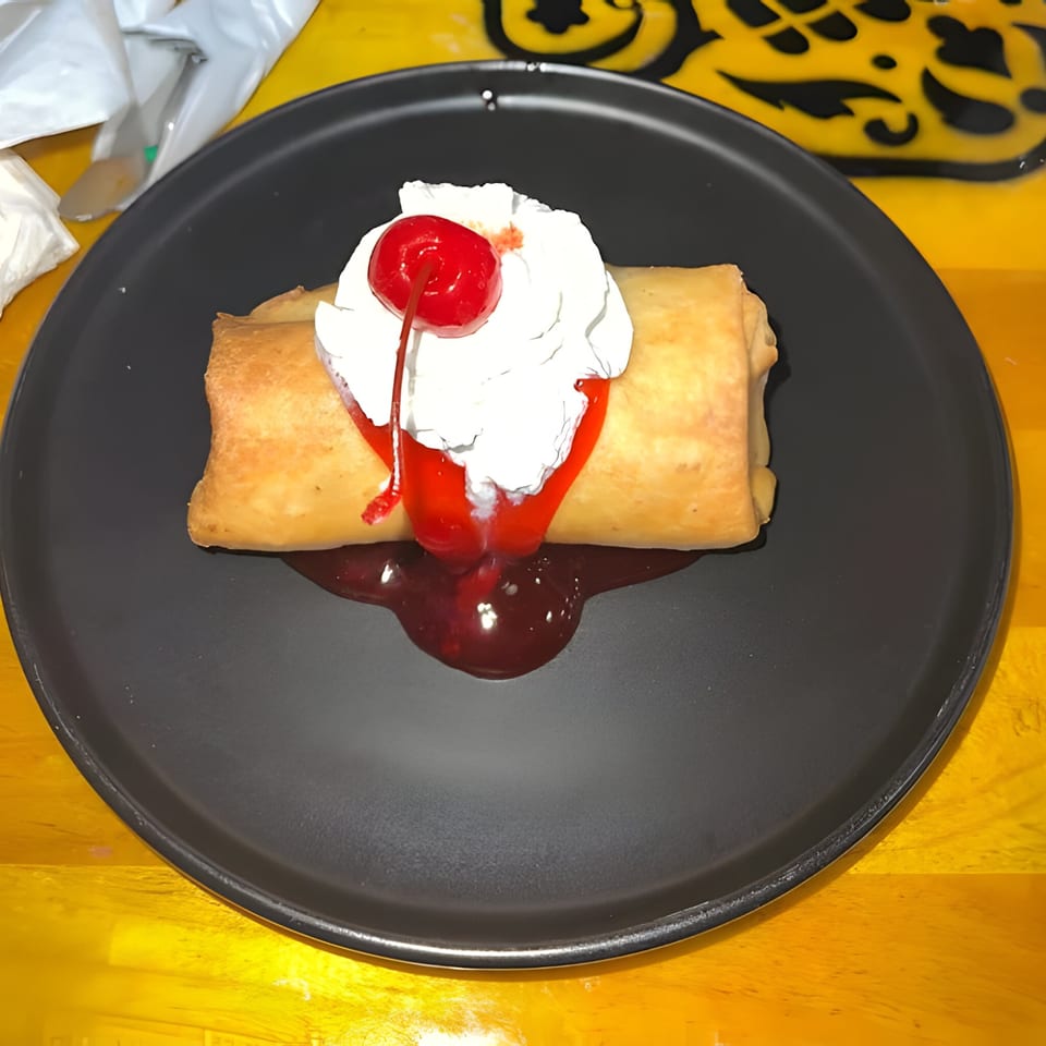 Golden-fried Mexican dessert, possibly a chimichanga, topped with whipped cream, cherry, and strawberry syrup.