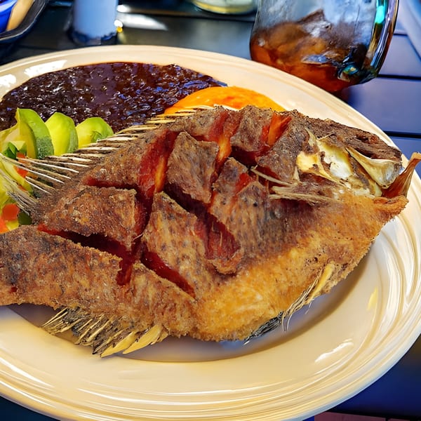 A whole fried fish with black beans, avocado slices, and orange sauce on a white plate.