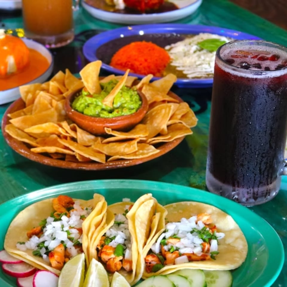 A vibrant Mexican feast with tacos, guacamole, chips, rice, and a dark beverage on a green table.