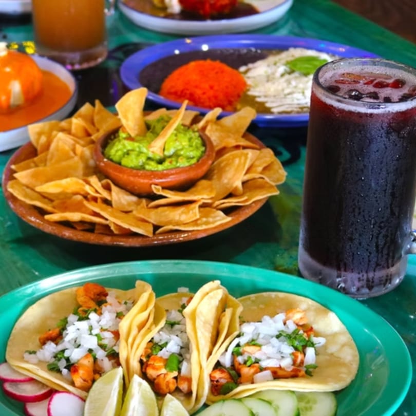 A vibrant Mexican feast with tacos, guacamole, chips, rice, and a dark beverage on a green table.